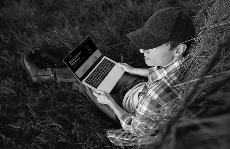Junger Landwirt sitzt im Gras, am Heuballen angelehnt, Laptop auf dem Schoß